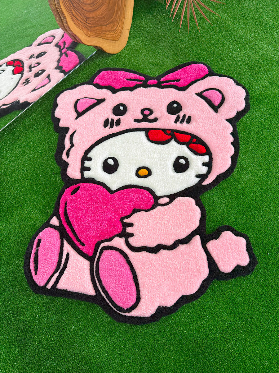 Pembe ayıcık kostümü giymiş ve elinde büyük pembe bir kalp tutan el yapımı Hello Kitty Pink Edition tufting halı; yumuşak dokusuyla öne çıkan, çocuk odaları için ideal özel tasarım ayıcıklı çocuk halısı modeli.