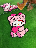 Yeşil çim zemin üzerinde, yanındaki doğal ahşap kütük dekoru ve arkasındaki aynadan yansıması görülen, pembe ayıcık kostümlü ve kalpli Hello Kitty Pink Edition el yapımı tufting halı konsept çekimi.