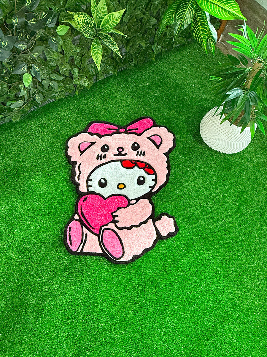 Yeşil çim zemin üzerinde, etrafında yapraklar ve beyaz dekoratif saksı ile sergilenen, pembe ayıcık kostümlü ve kalpli Hello Kitty Pink Edition el yapımı tufting halı konsept çekimi.