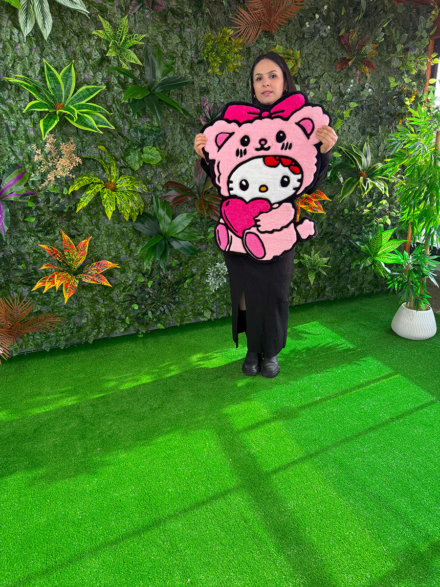 Yeşil bitki duvarı önünde, kadın model tarafından elde tutularak gerçek boyutları sergilenen, pembe ayıcık kostümlü ve kalpli Hello Kitty Pink Edition el yapımı tufting halı.