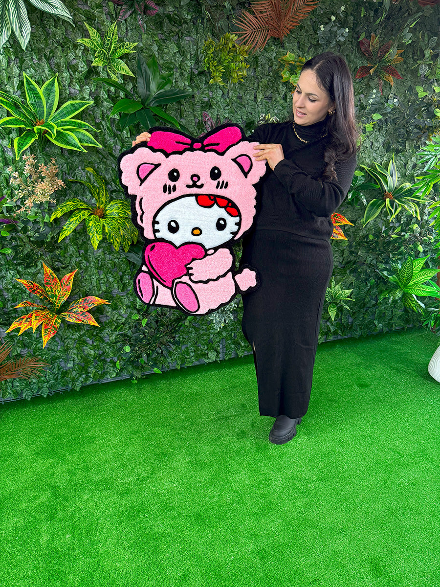 Yeşil bitki duvarı önünde, kadın modelin elinde farklı bir açıyla sergilenen, pembe ayıcık kostümlü ve kalpli Hello Kitty Pink Edition el yapımı tufting halı.