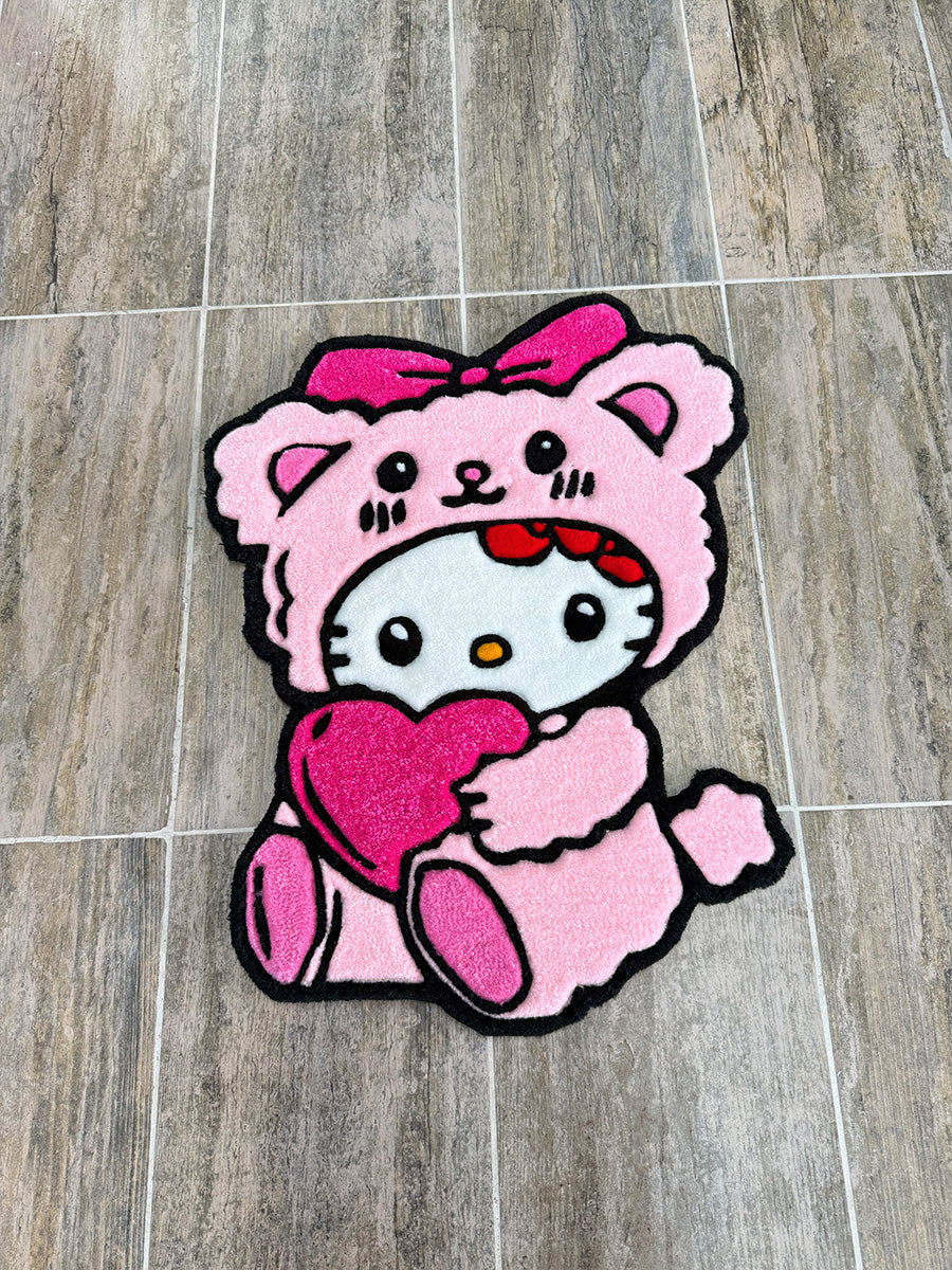 Pembe ayıcık kostümü giymiş ve elinde kalp tutan Hello Kitty Pink Edition el yapımı tufting halı modelinin tam boy üstten görünümü. Çocuk odası dekorasyonuna uygun, kalın dokulu, siyah dış hatlara ve canlı renklere sahip özel tasarım pembe Hello Kitty tufting halısı.