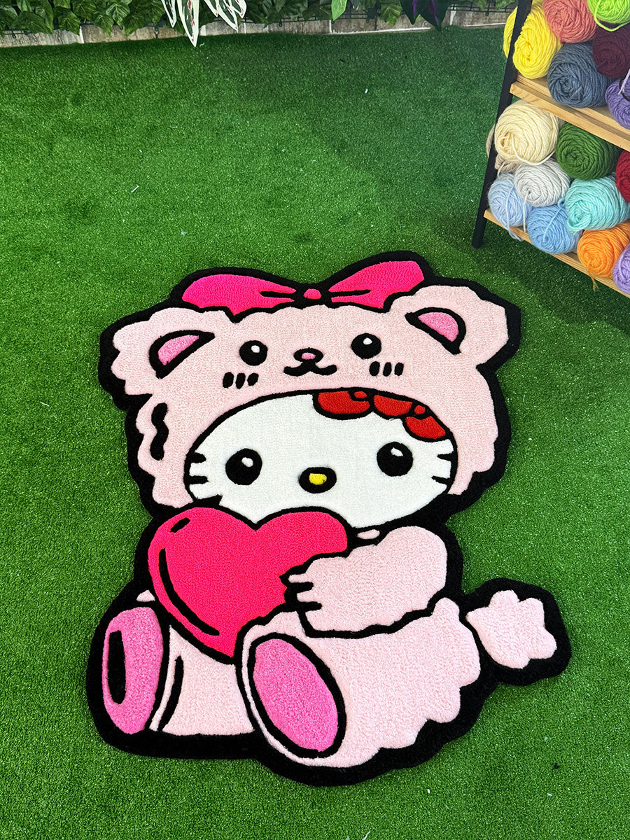 Renkli yumakların yanında, çim zemin üzerinde sergilenen pembe ayıcık kostümlü ve kalpli Hello Kitty Pink Edition el yapımı tufting halı tam görünümü.