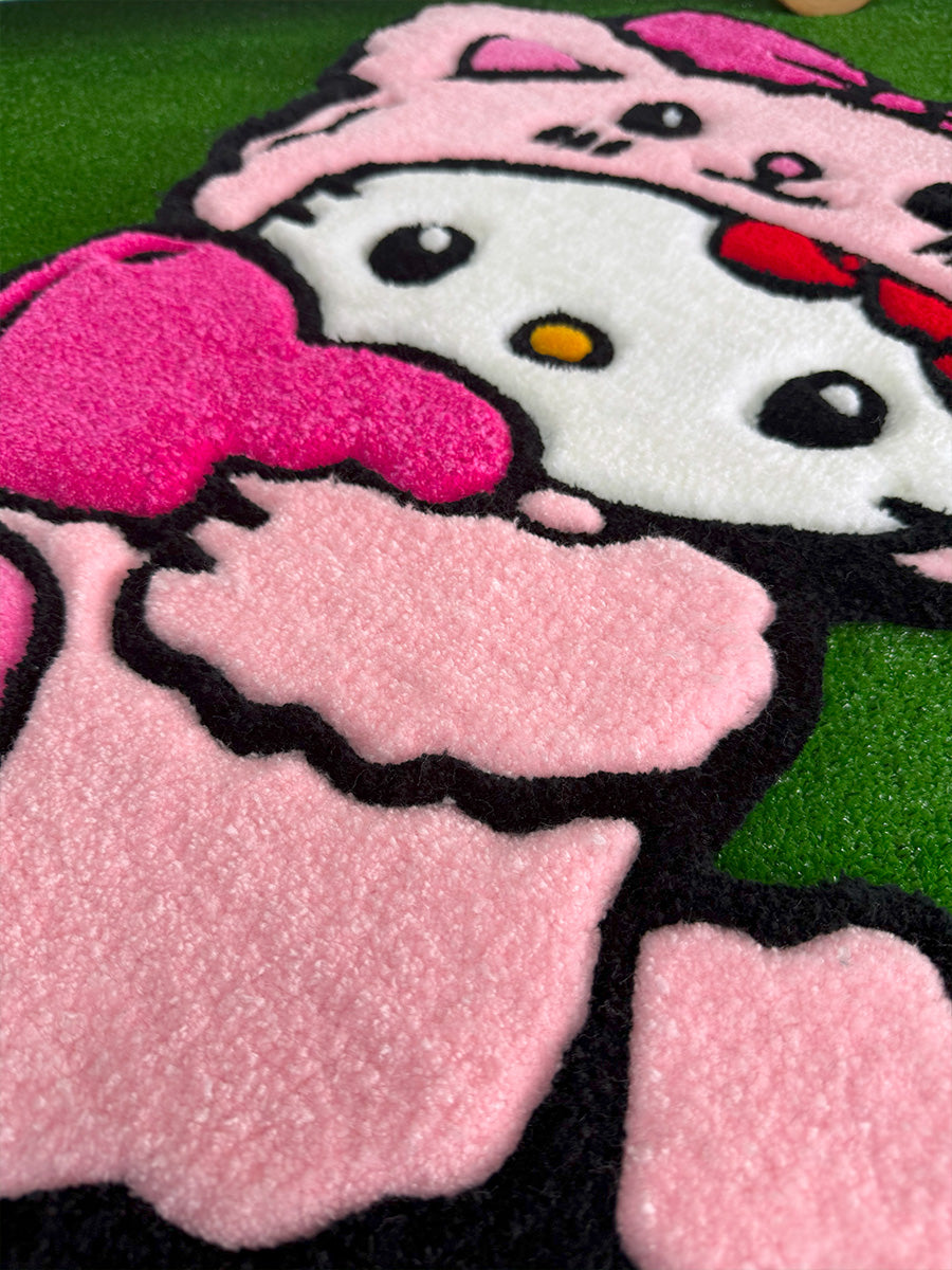 Hello Kitty Pink Edition el yapımı tufting halı modelinin yakından çekilmiş detay görseli. Bu açıda, pembe ayıcık kostümünün puf puf iplik dokusu, tuttuğu pembe kalbin canlı renkleri ve tasarıma üç boyutlu bir görünüm kazandıran özenle tıraşlanmış kalın siyah kontur çizgileri net bir şekilde ön plana çıkmaktadır.