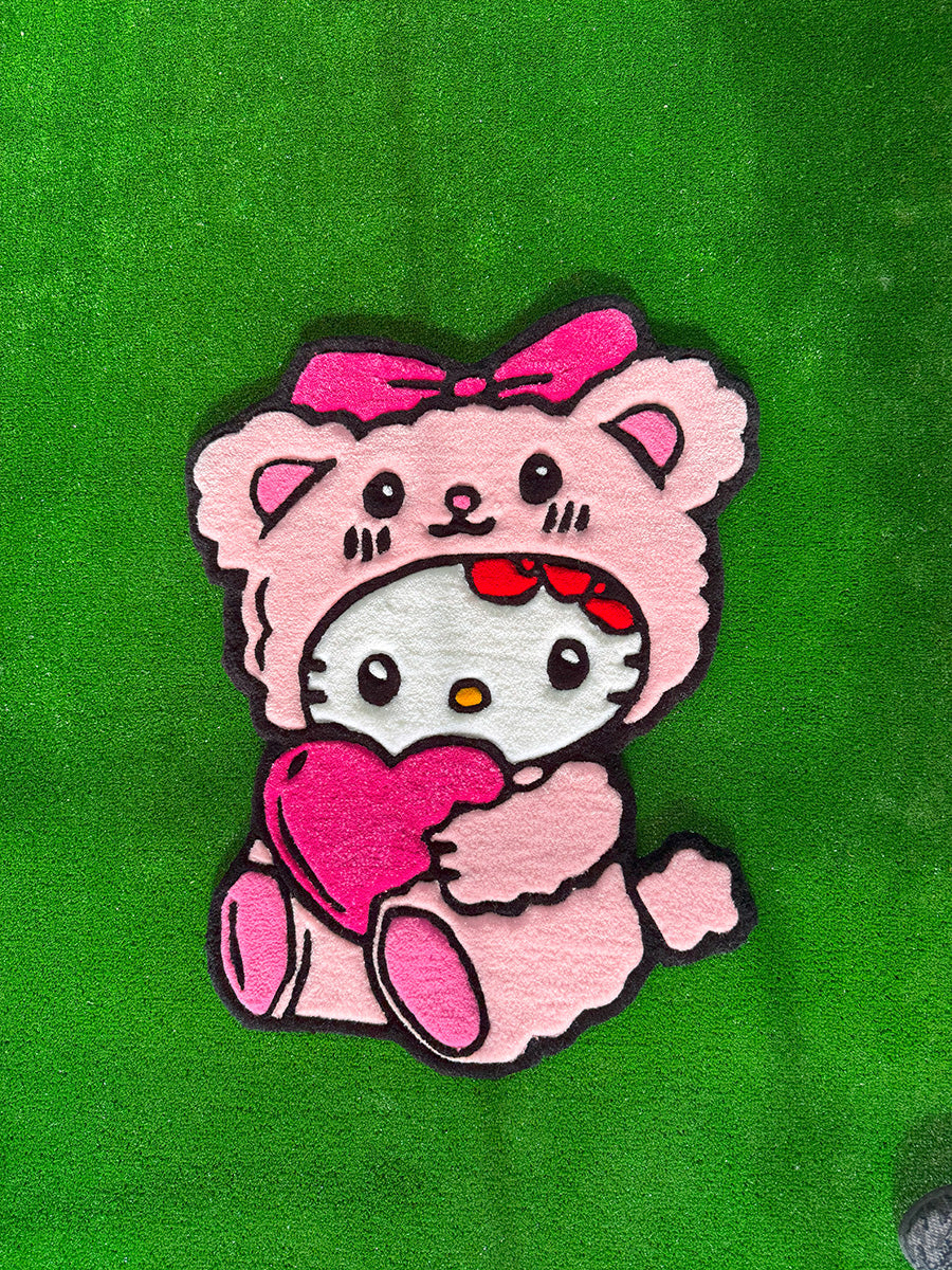 Yeşil çim zemin üzerinde tek başına sergilenen, pembe ayıcık kostümü giymiş ve kalp tutan Hello Kitty Pink Edition el yapımı tufting halının sade ve net tam boy görünümü.