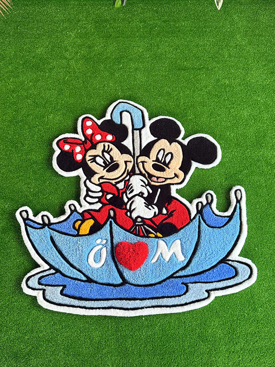 Üzerinde kişiselleştirilmiş harfler ve belirgin kırmızı bir kalp bulunan mavi şemsiyeli Mickey ve Minnie Mouse çocuk halısı tasarımının tam üstten çekilmiş net ve düz görünümü. Siyah kalın konturlarla sınırları keskinleştirilmiş, el işçiliğiyle üretilen tufting dekoratif çocuk odası halısı.