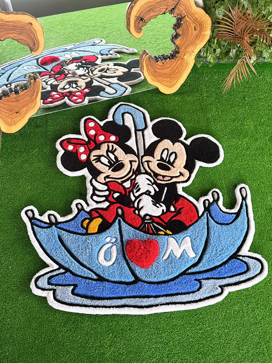 Mavi bir şemsiye altında neşeyle birbirine sarılan Mickey ve Minnie Mouse figürlerinin işlendiği, kırmızı kalp detaylı el yapımı tufting halı. Canlı renklere ve yumuşak dokuya sahip bu dekoratif çocuk odası halısı modelinin, detayları öne çıkaran yakından çekilmiş açılı görünümü.